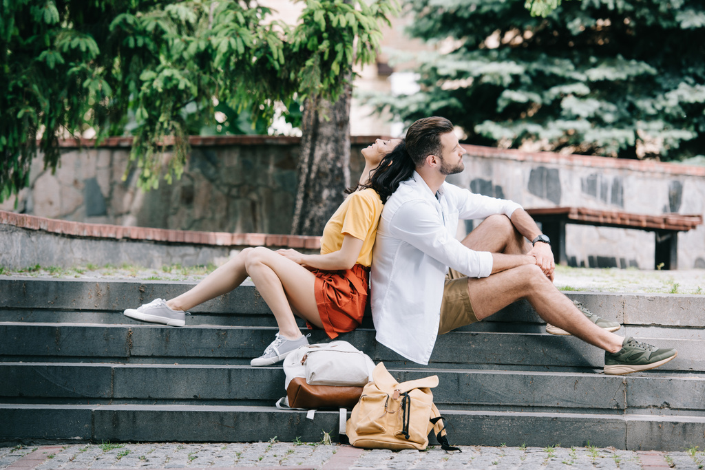 side view of bearded man and cheerful woman sitting on stairs near backpacks  - Photo, Image