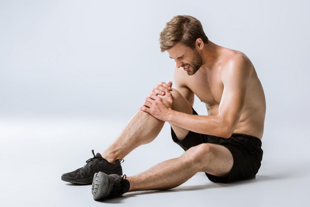 бородатый спортсмен без рубашки с болью в коленях на сером
 - Фото, изображение