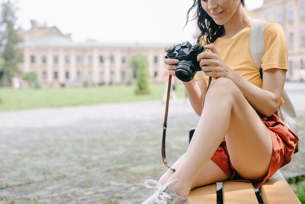 vista ritagliata di giovane donna in possesso di fotocamera digitale mentre seduto sulla panchina vicino all'università
  - Foto, immagini