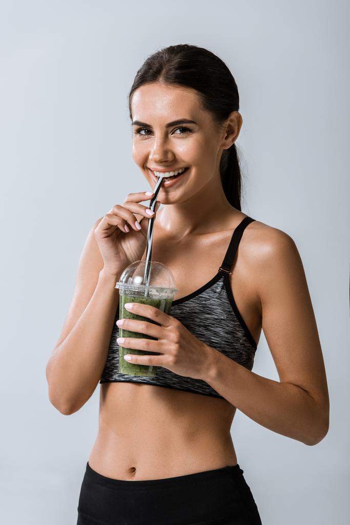 attractive smiling sportswoman drinking smoothie isolated on grey - Photo, Image