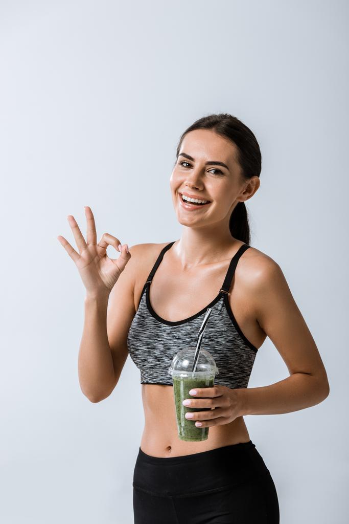 smiling sportswoman holding smoothie and showing okay sign isolated on grey - Photo, Image