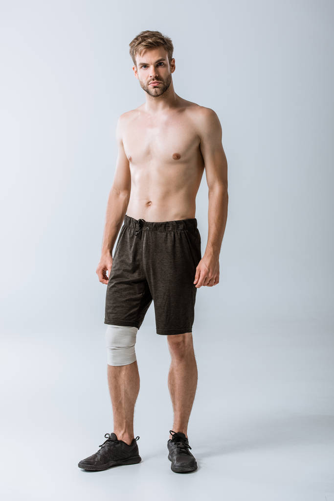 Ganzkörperansicht des hemdlosen Sportlers mit elastischem Verband am Knie auf grau - Foto, Bild
