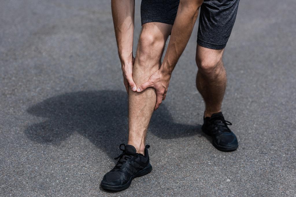 partial view of sportsman in black sneakers with leg pain on street - Photo, Image