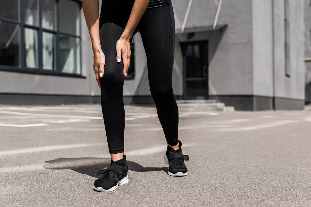 περικομμένη όψη του αθλητικού αθλητισμού σε μαύρα αθλητικά παπούτσια με πόνο στο γόνατο στο δρόμο - Φωτογραφία, εικόνα