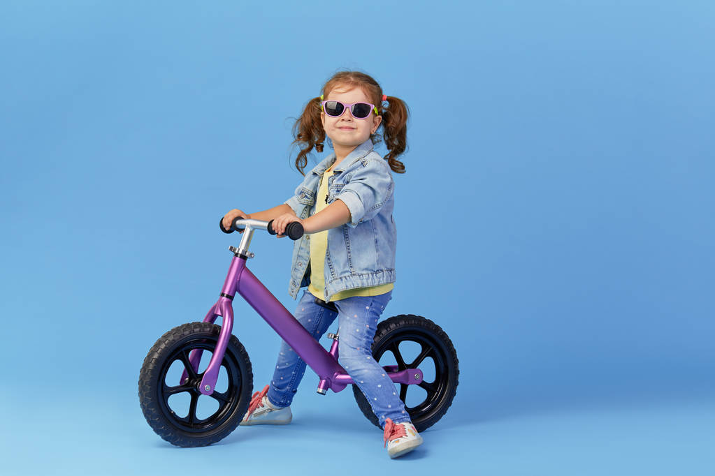 Petite fille apprend à garder l'équilibre tout en faisant du vélo. Enfant élégant dans des lunettes de soleil isolées sur fond bleu
 - Photo, image