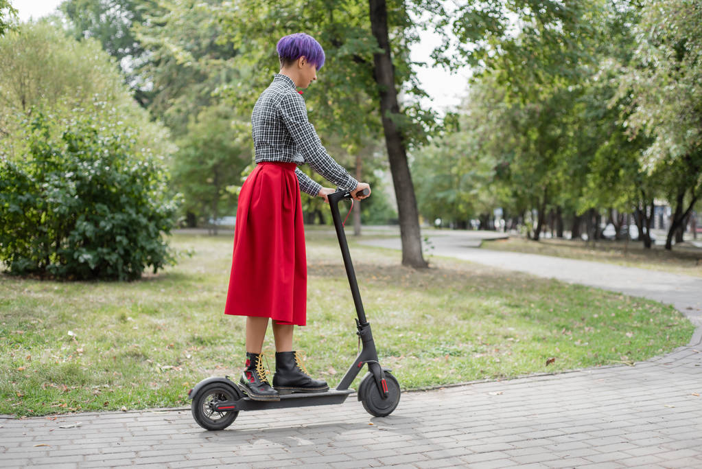 Una joven con el pelo morado monta una scooter eléctrica en un parque. Una chica elegante con un templo afeitado en una camisa a cuadros, una falda roja larga y una corbata de lazo está montando alrededor de la ciudad en un dispositivo moderno
.  - Foto, imagen