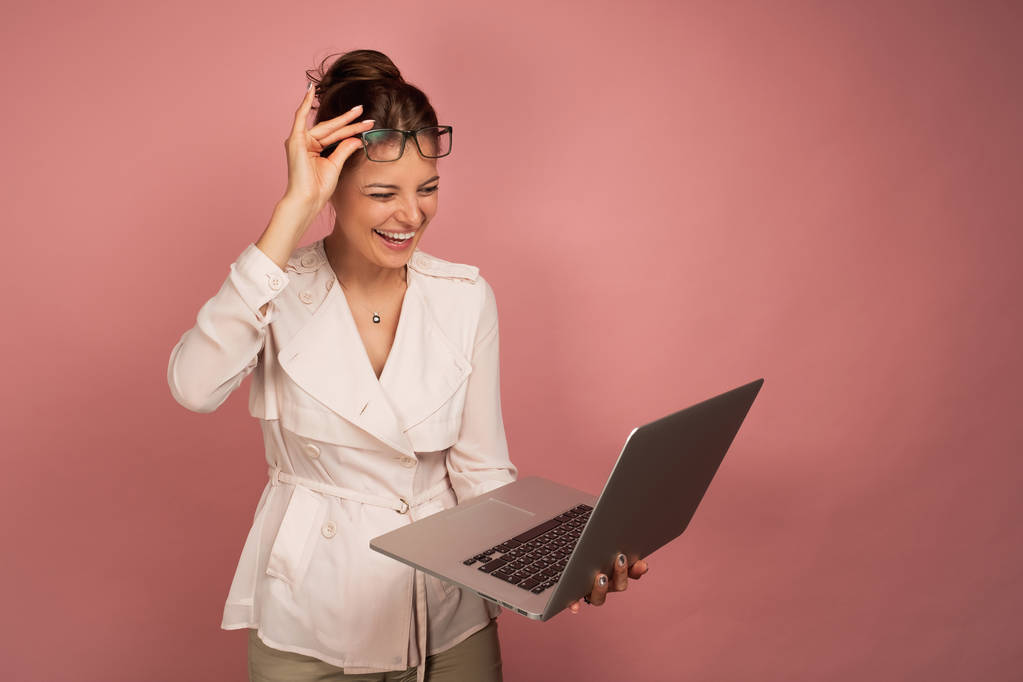 A girl in a light jacket stands on a pink background and holds a laptop in her hand laughing and raising glasses on her forehead. - Photo, Image