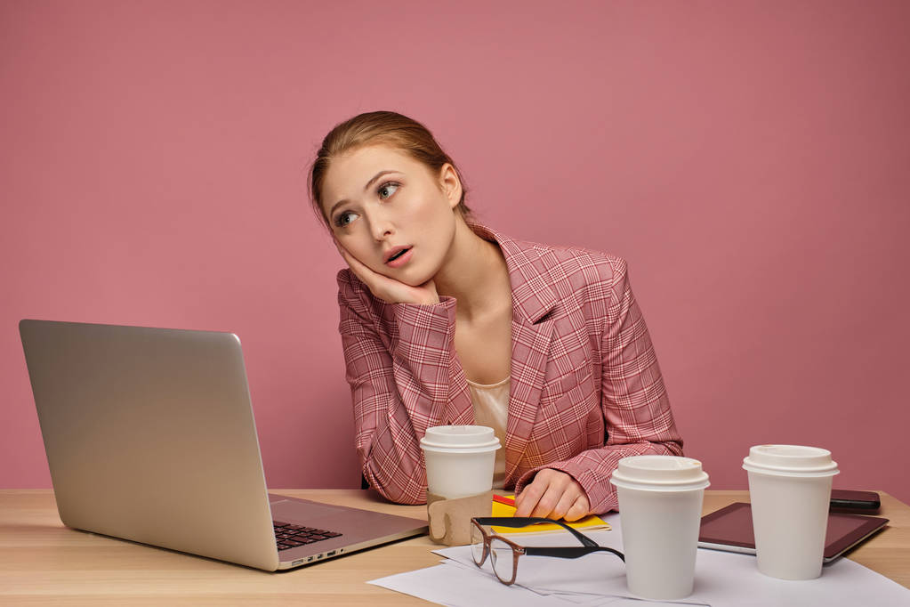 Ragazza rossa in un abito su uno sfondo rosa si siede davanti a un computer portatile e sostiene stancamente la testa con la mano
. - Foto, immagini