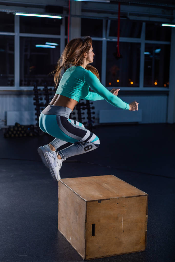 Fit young woman doing box jumping on dark background with copy space. High jump on one foot on a box. Beautiful athlete performs a circular workout. Squatting . - Photo, Image