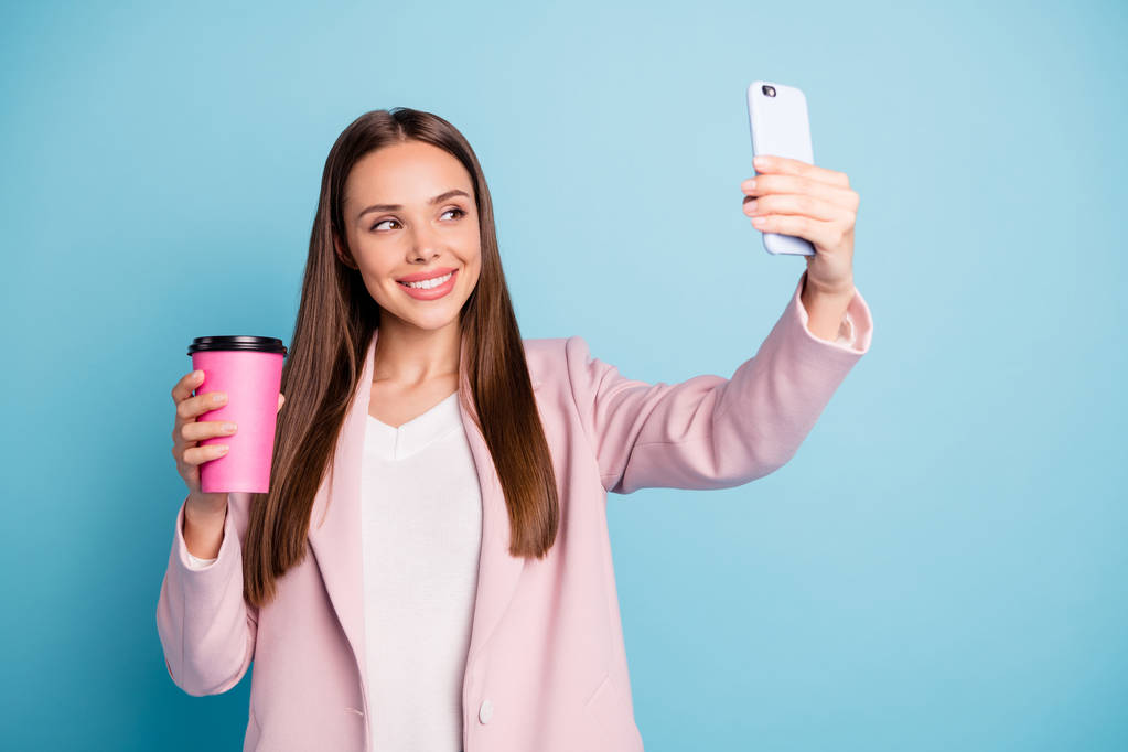 Portrait of cute cheerful lady using her cellphone making selfie holding mug with hot beverage wearing pink coat isolated over blue color background - Photo, Image