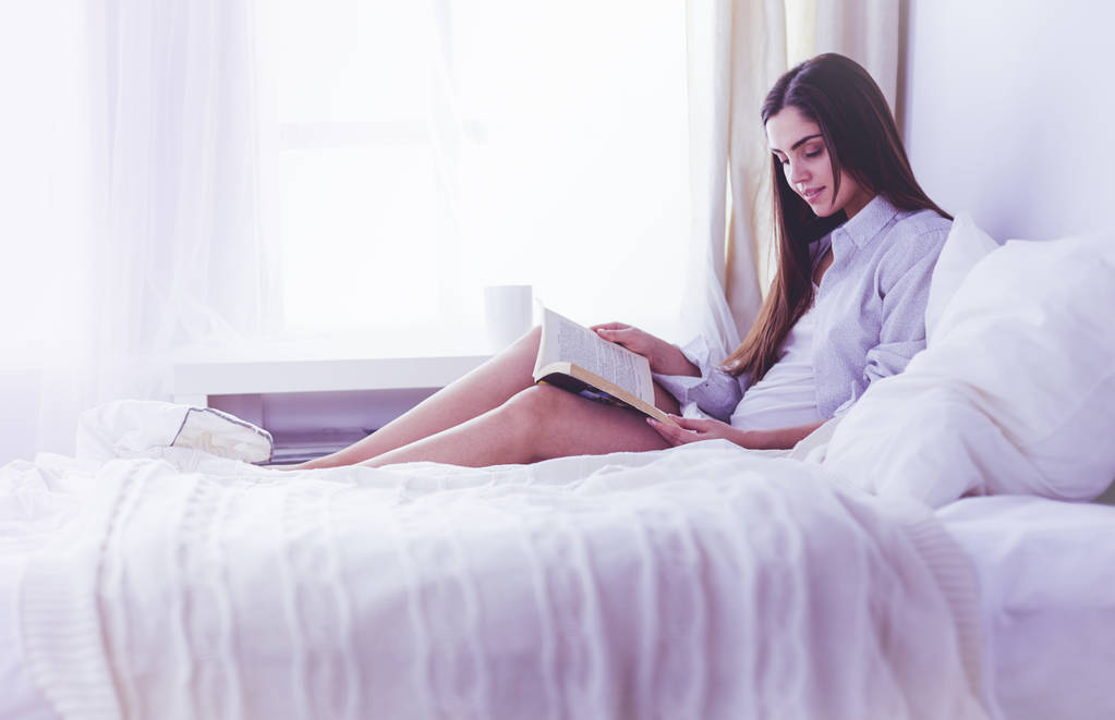 Portré egy nőről, aki tablettát használ és kávét iszik, miközben reggel az ágyon ül. - Fotó, kép