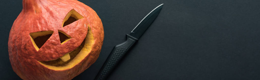 Panoramaaufnahme eines Halloween-Kürbisses in der Nähe eines Messers auf schwarzem Hintergrund - Foto, Bild
