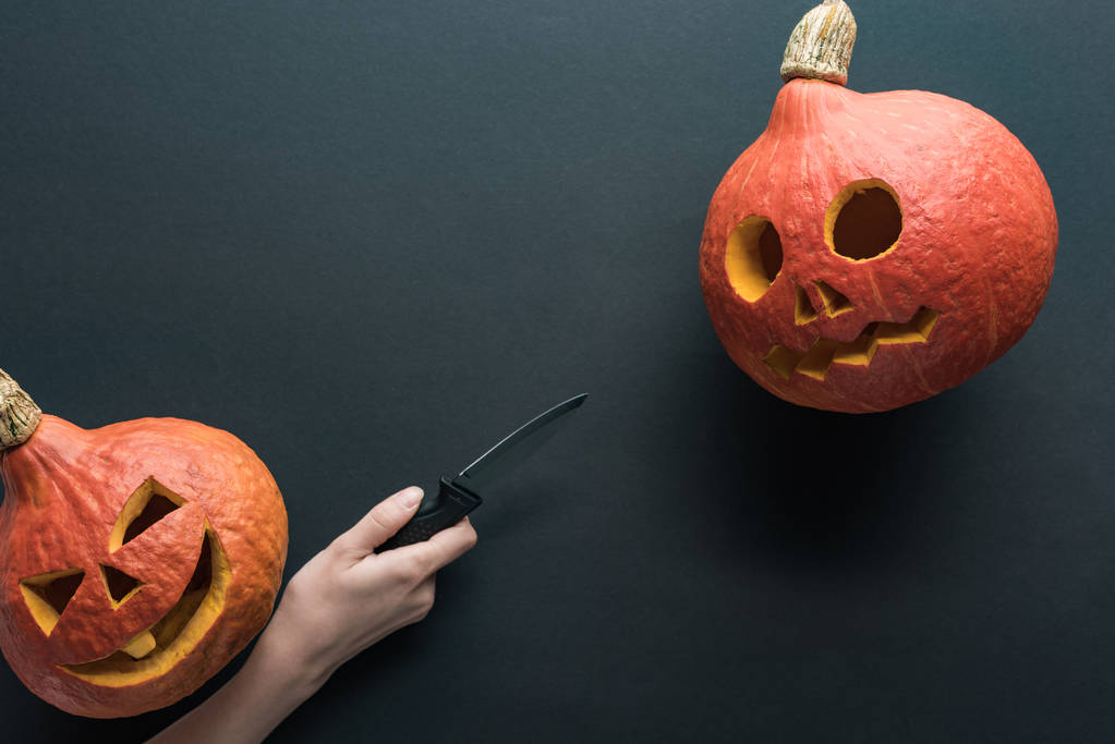 partial view of woman holding knife near carved spooky Halloween pumpkins on black background - Photo, Image