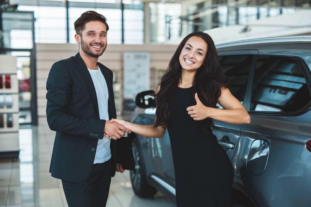 Красивый менеджер мужчина и красивая брюнетка клиентка пожимает руку после успешной сделки в дилерском центре женщина показывает, как стоит рядом с совершенно новой машиной
 - Фото, изображение