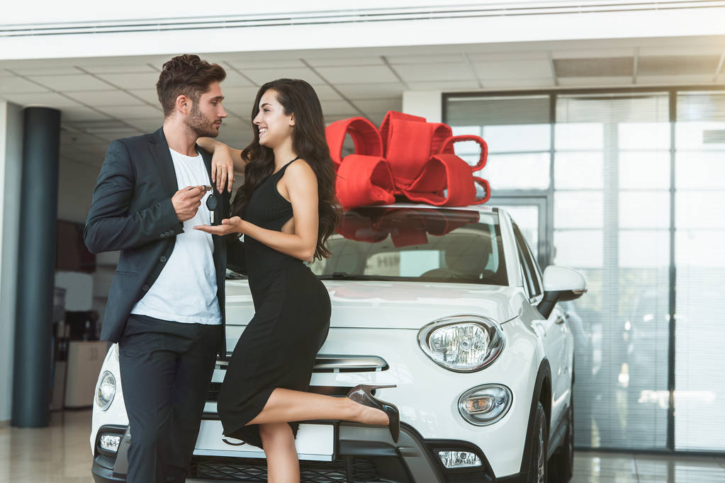 junger gutaussehender Mann übergibt Schlüssel von nagelneuem Auto, das er seiner schönen Frau präsentierte, die im Autohaus steht und glücklich und zufrieden aussieht - Foto, Bild