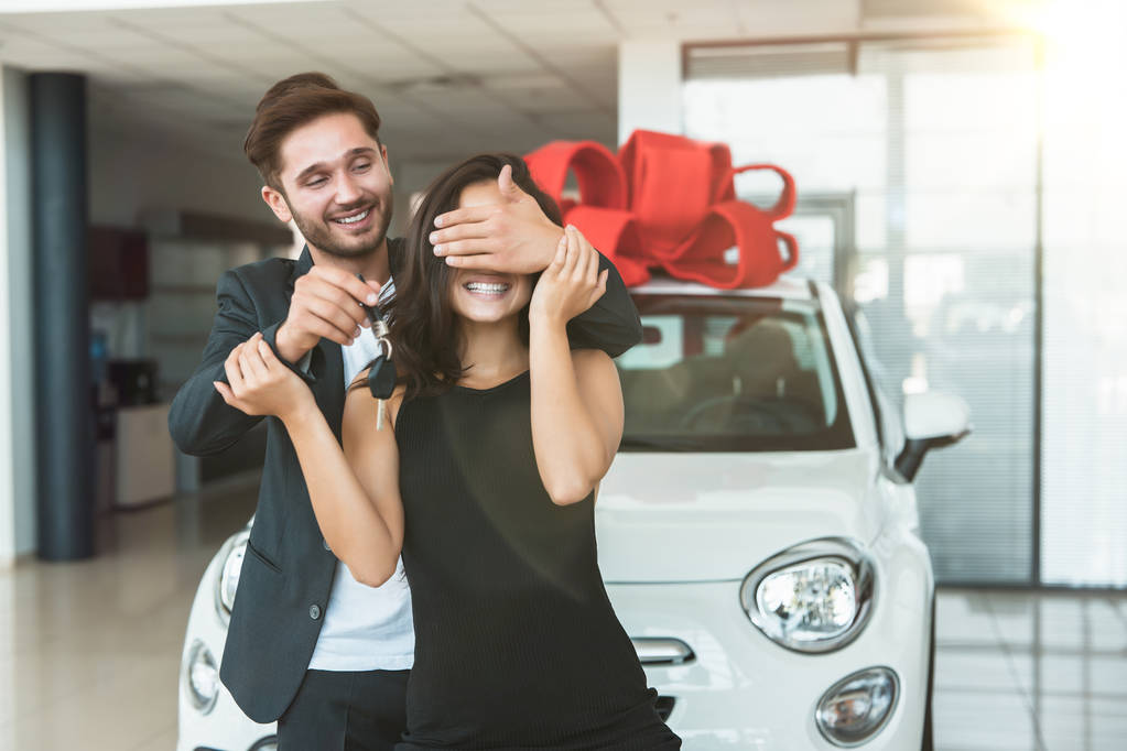 jonge knappe man maakt verrassing voor zijn mooie vrouw staande in dealer Center kopen gloednieuwe auto voor heden - Foto, afbeelding