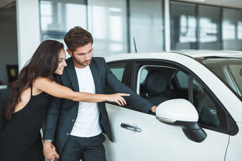 hermosa pareja joven hombre y mujer elegir un coche nuevo en el centro de concesionarios buscando emocionado
 - Foto, Imagen
