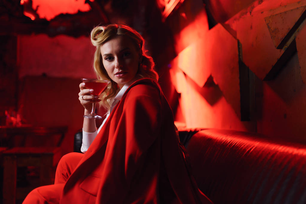Imagem de loira séria em casaco vermelho olhando para a câmera com coquetel na mão em boate
 - Foto, Imagem