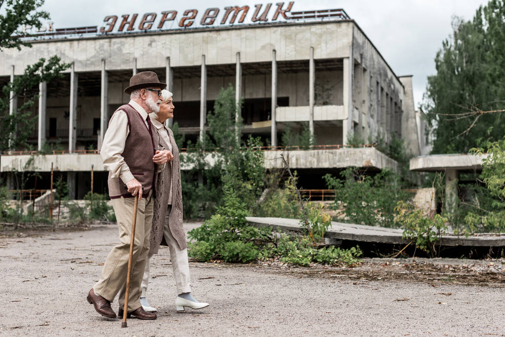 pripjat, Ukraine - 15. August 2019: Rentnerehepaar spaziert in Tschernobyl in der Nähe eines Gebäudes mit energiegeladenen Schriftzügen  - Foto, Bild