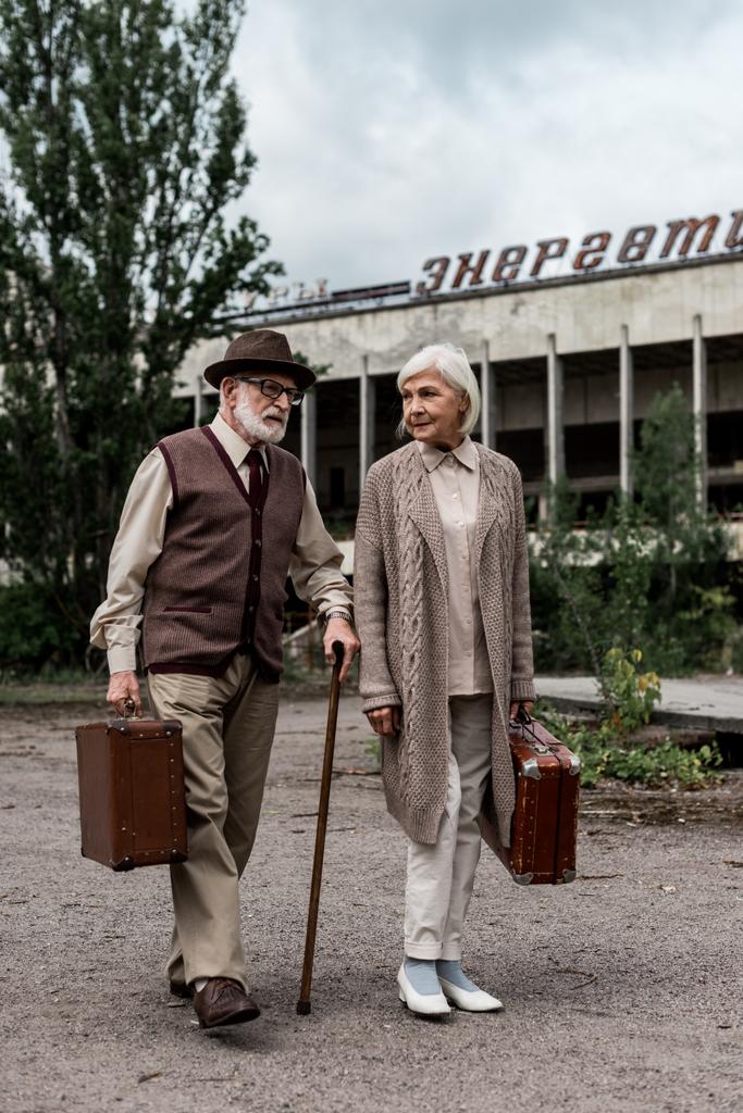Pripyat, Ουκρανία-15 Αυγούστου 2019: συνταξιούχος ζευγάρι με βαλίτσες κοντά στο κτίριο με ενεργειακά γράμματα στο Τσερνομπίλ  - Φωτογραφία, εικόνα