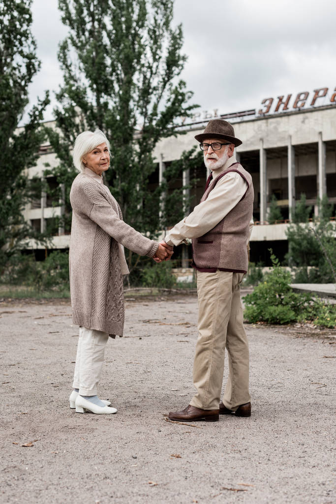 Pripyat, Ουκρανία-15 Αυγούστου 2019: ζευγάρι ηλικιωμένων που κρατά τα χέρια κοντά στο κτίριο με ενεργητικά γράμματα στο Τσερνομπίλ  - Φωτογραφία, εικόνα