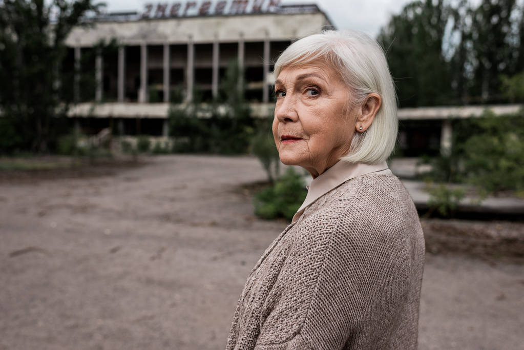 PRIPYAT, UCRÂNIA - AGOSTO 15, 2019: mulher sênior olhando para a câmera perto do edifício em chernobyl
  - Foto, Imagem