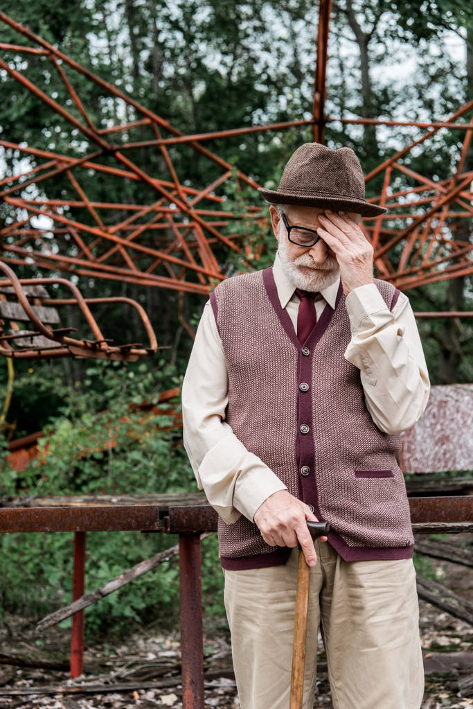 ПРИПЯТ, УКРАИНА - 15 августа 2019 года: бородатый пенсионер в шляпе и очках стоит с тростью рядом с поврежденной каруселью
  - Фото, изображение
