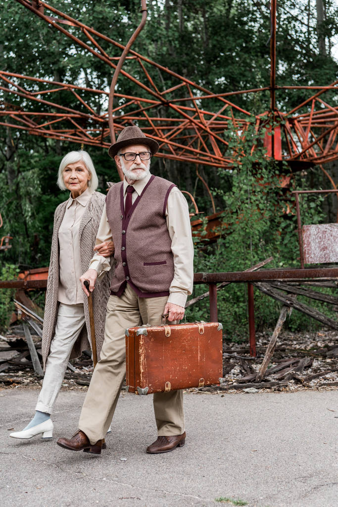 Prypyat, Ukraina-15 sierpnia 2019: starszy mężczyzna w okularach trzyma walizkę podczas chodzenia z żoną w pobliżu opuszczonej karuzeli  - Zdjęcie, obraz