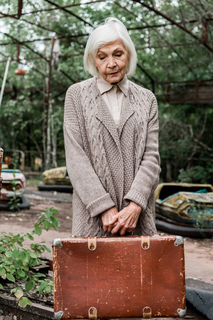 ПРИПЯТ, УКРАИНА - 15 августа 2019 года: грустная пожилая женщина с чемоданом в парке развлечений
  - Фото, изображение