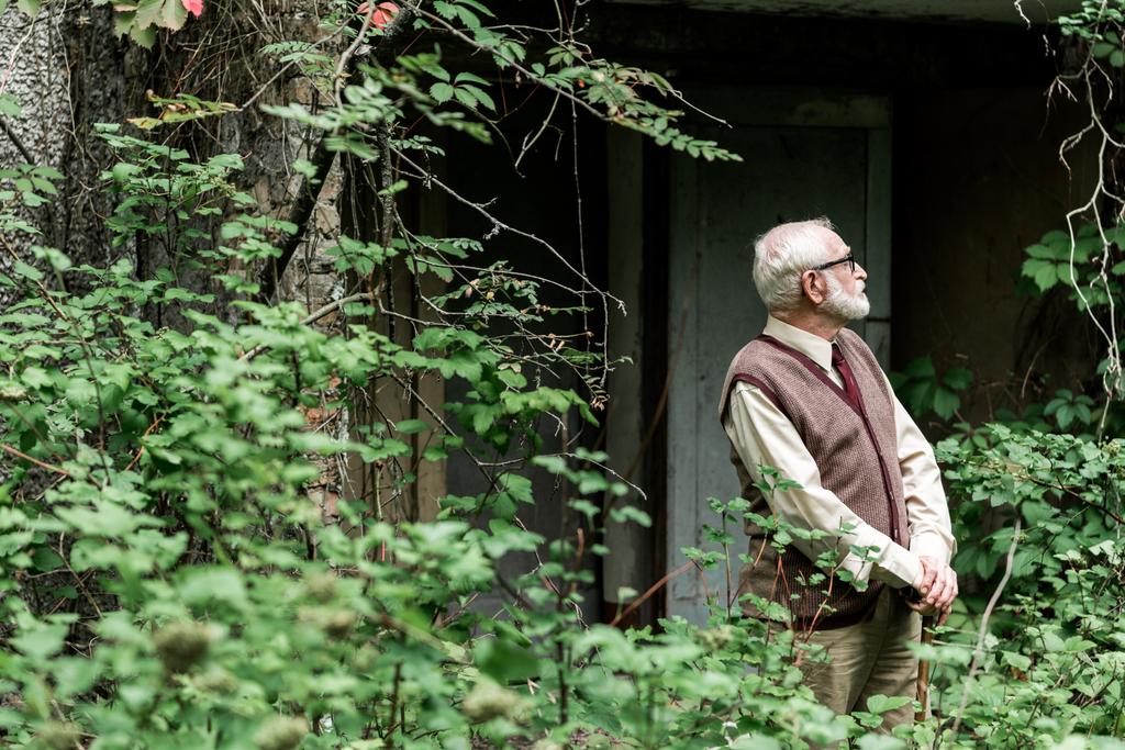 επιλεκτική εστίαση του συνταξιούχου άντρα σε γυαλιά που στέκονται με το μπαστούνι κοντά στο σπίτι  - Φωτογραφία, εικόνα