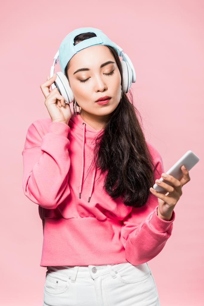 attraktive asiatische Frau in Pullover und Mütze hört Musik und hält ihr Smartphone isoliert auf rosa  - Foto, Bild