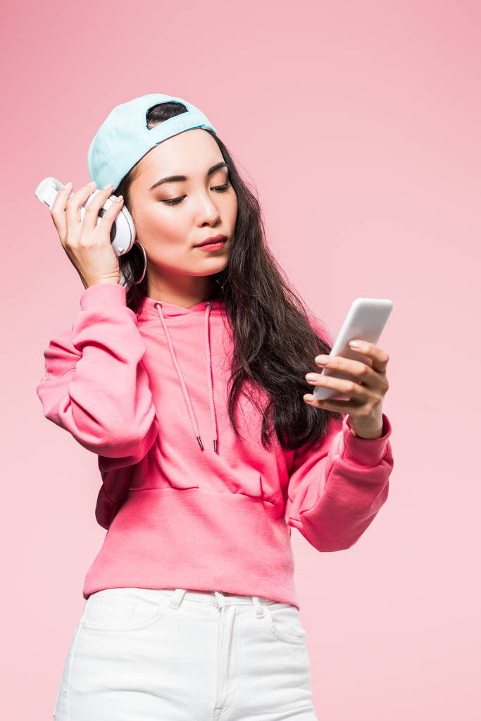 attraente asiatico donna in pullover e cap ascolto musica e tenendo smartphone isolato su rosa
  - Foto, immagini