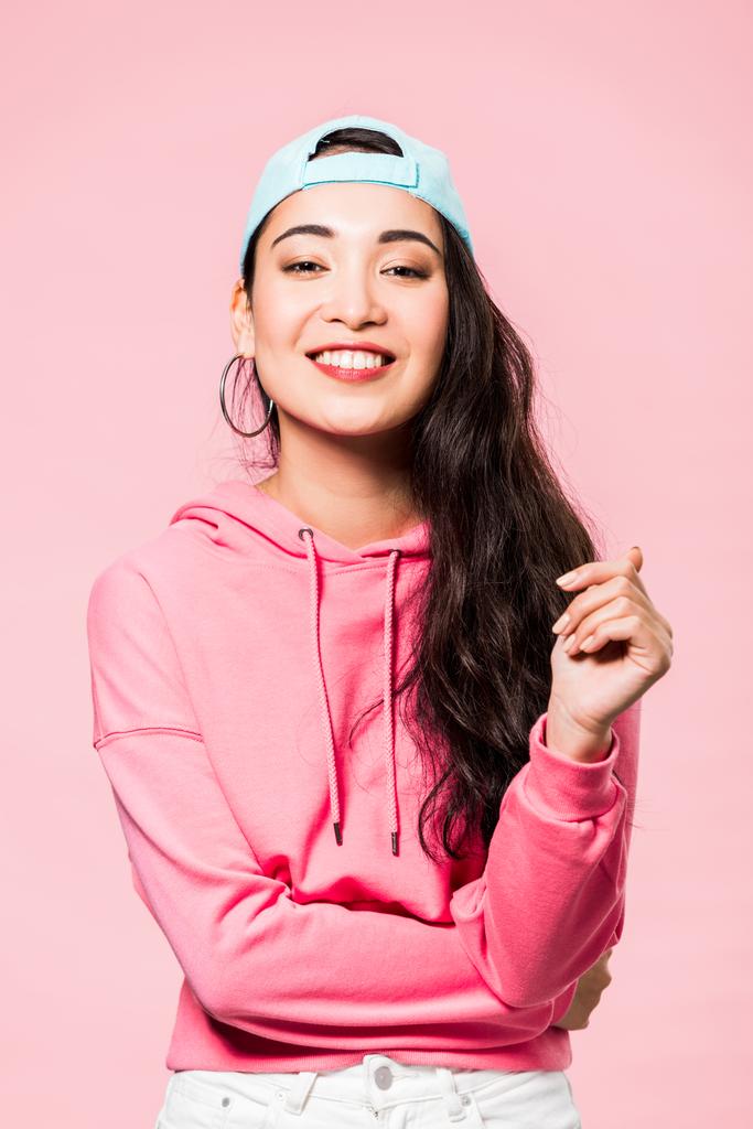 attractive asian woman in pink pullover and cap smiling isolated on pink  - Photo, Image