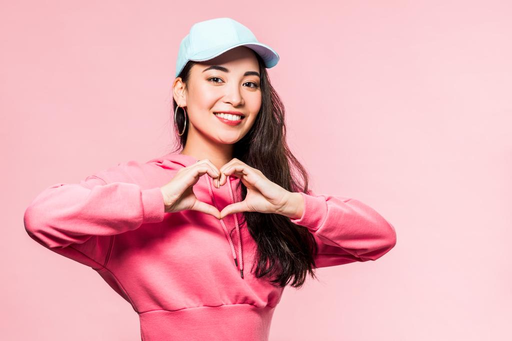 attraente donna asiatica in pullover rosa e cappuccio sorridente e mostrando il simbolo del cuore isolato sul rosa
  - Foto, immagini