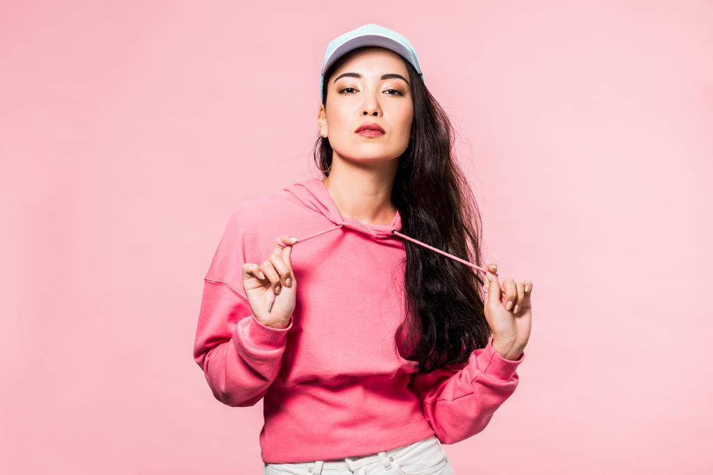 atractiva mujer asiática en rosa jersey y gorra mirando cámara aislada en rosa
  - Foto, Imagen