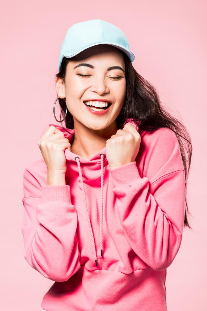 atraente asiático mulher com fechado olhos no rosa pulôver e cap sorrindo isolado no rosa
  - Foto, Imagem