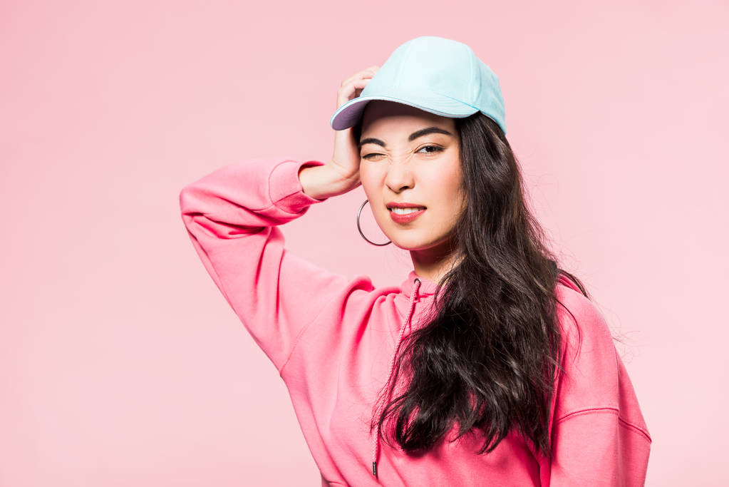 attractive asian woman in pink pullover and cap winking isolated on pink  - Photo, Image