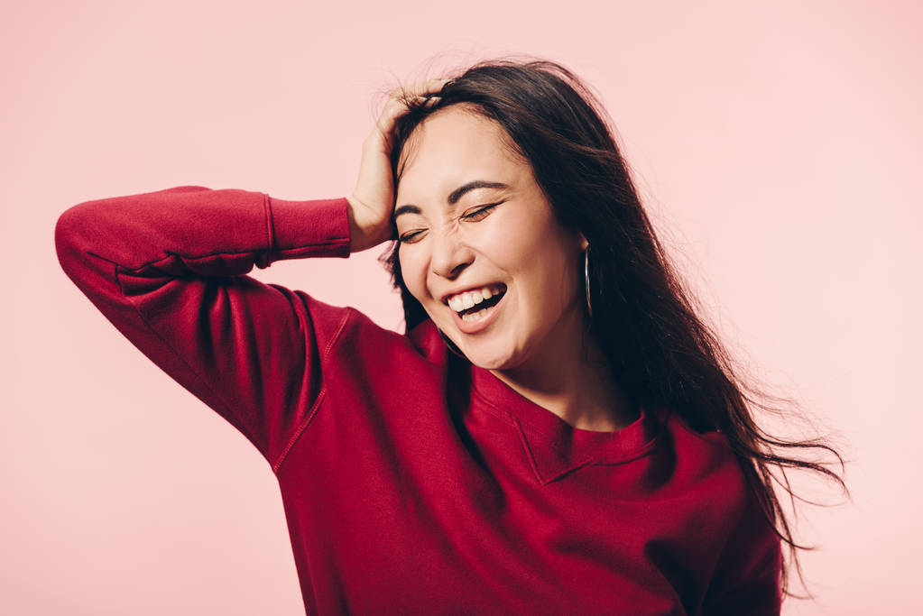 attractive asian woman in red sweater with closed eyes smiling isolated on pink  - Photo, Image