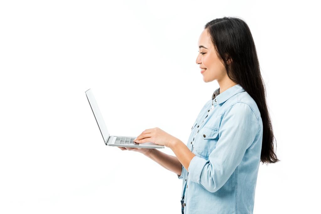πλαϊνή άποψη της ελκυστικής γυναίκας της Ασίας σε denim πουκάμισο χαμογελώντας και κρατώντας φορητό υπολογιστή απομονωμένο σε λευκό  - Φωτογραφία, εικόνα