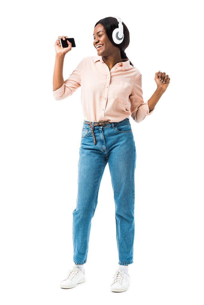 sonriente mujer afroamericana en camisa con auriculares sosteniendo teléfono inteligente y bailando aislado en blanco
  - Foto, imagen