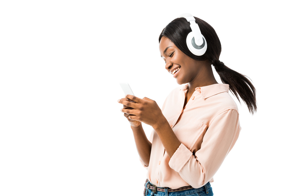 усміхнена афроамериканська жінка в сорочці з навушниками за допомогою смартфона ізольована на білому
  - Фото, зображення
