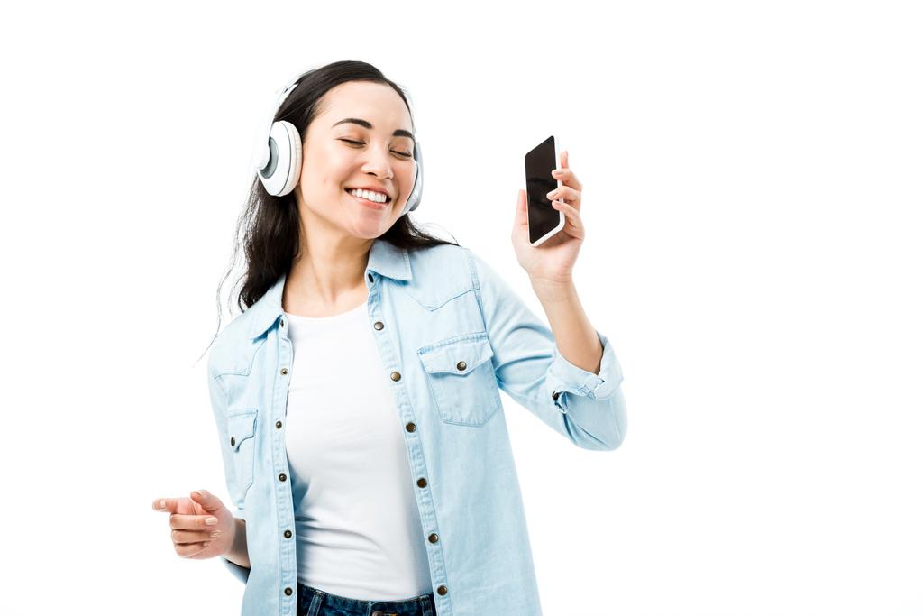 привлекательная и улыбающаяся азиатка в джинсовой рубашке слушает музыку и держит смартфон изолированным на белом
  - Фото, изображение