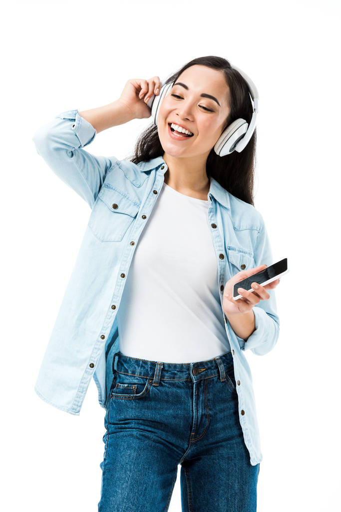 attractive and smiling asian woman in denim shirt listening music and holding smartphone isolated on white  - Photo, Image