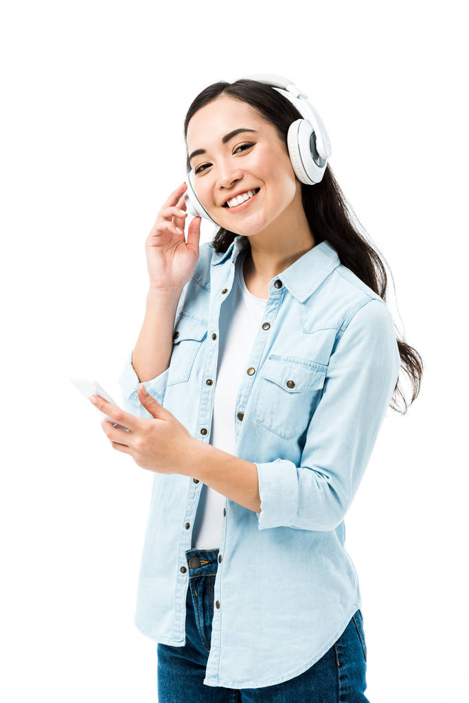 Kot tişörtlü çekici ve gülümseyen Asyalı kadın müzik dinliyor ve elinde beyaz bir telefon tutuyor.  - Fotoğraf, Görsel
