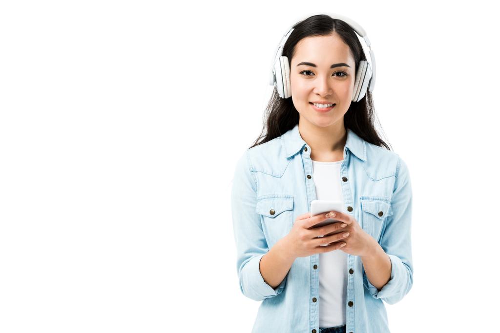 houkutteleva ja hymyilevä aasialainen nainen denim-paidassa kuuntelemassa musiikkia ja pitämässä älypuhelinta eristyksissä valkoisella
  - Valokuva, kuva