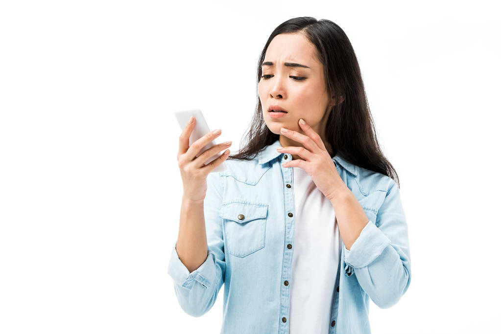 attractive and shocked asian woman in denim shirt holding smartphone isolated on white  - Photo, Image
