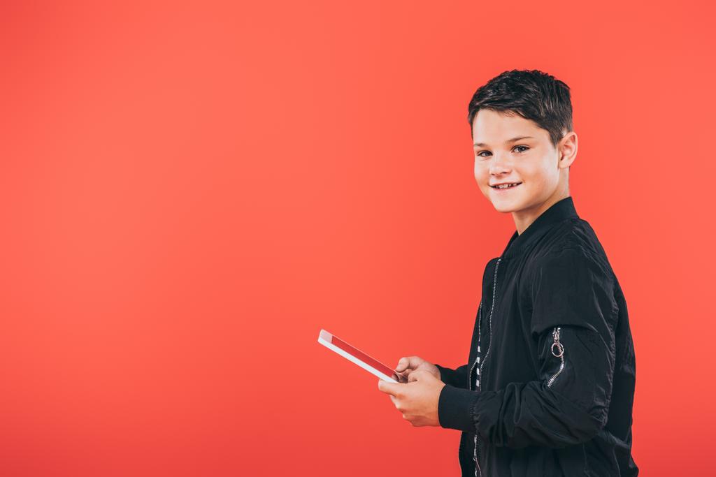 χαμογελαστή παιδί στο μπουφάν χρησιμοποιώντας ψηφιακό δισκίο απομονώνεται σε κόκκινο - Φωτογραφία, εικόνα
