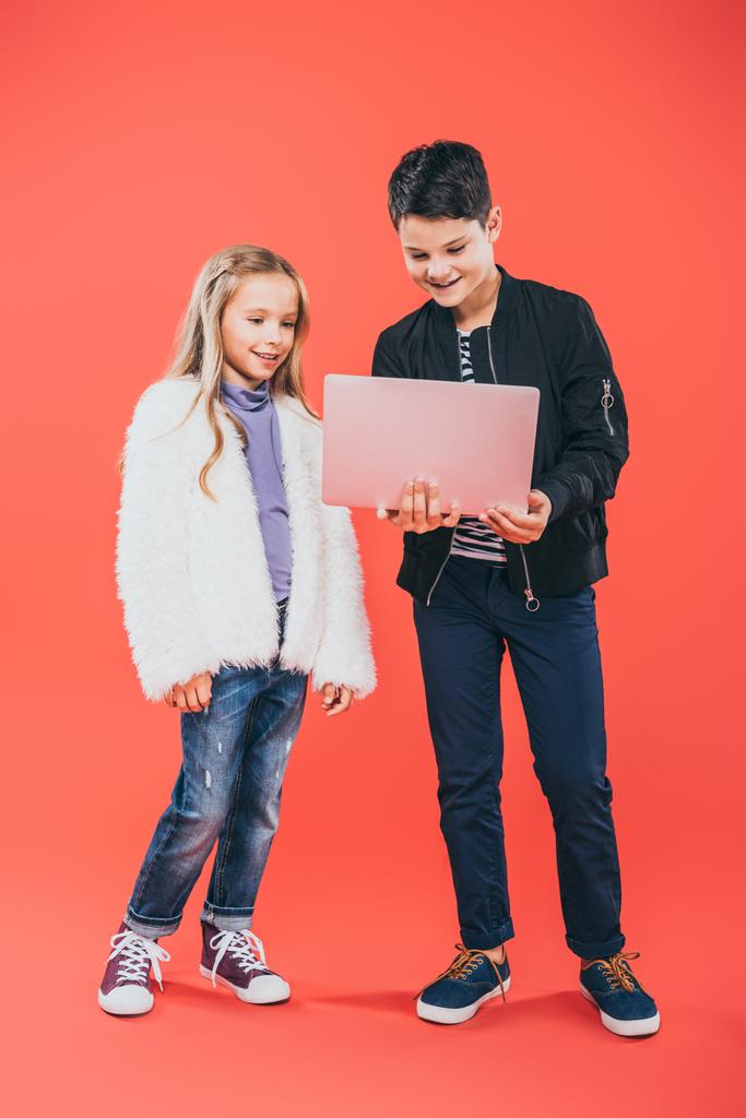 volledige lengte weergave van lachende kinderen met behulp van laptop op rood - Foto, afbeelding