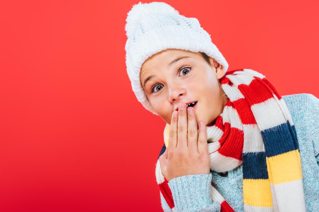 surprised kid in hat and scarf covering mouth with hand isolated on red - Photo, Image