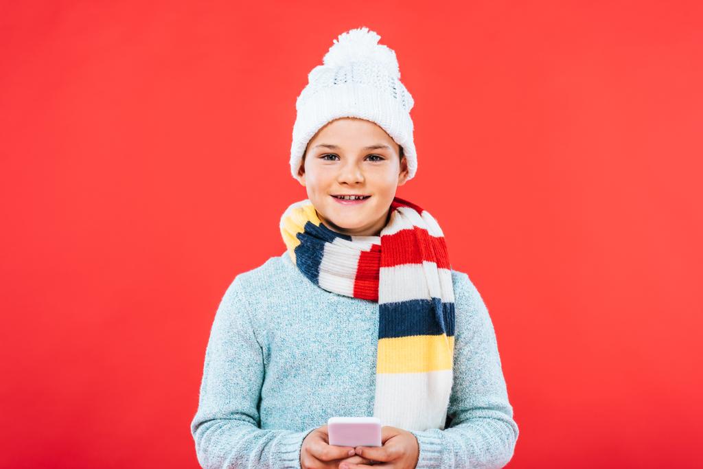 front view of smiling kid in hat and scarf using smartphone isolated on red - Photo, Image